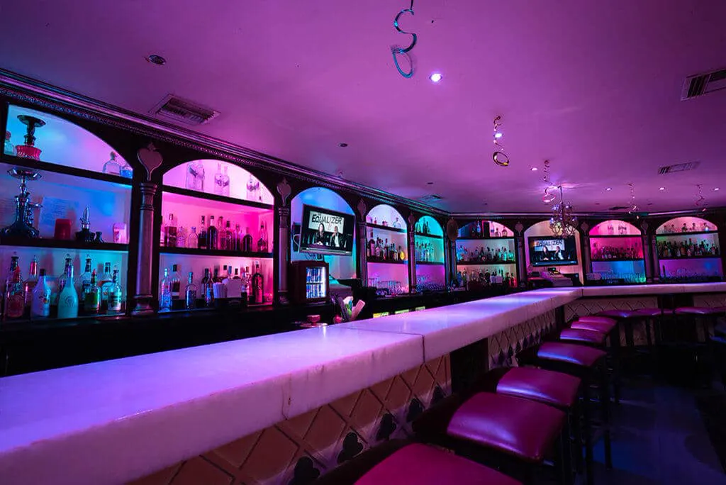 Long bar at Bella's Cabaret lit in pink and purple, with stools and backlit liquor shelves